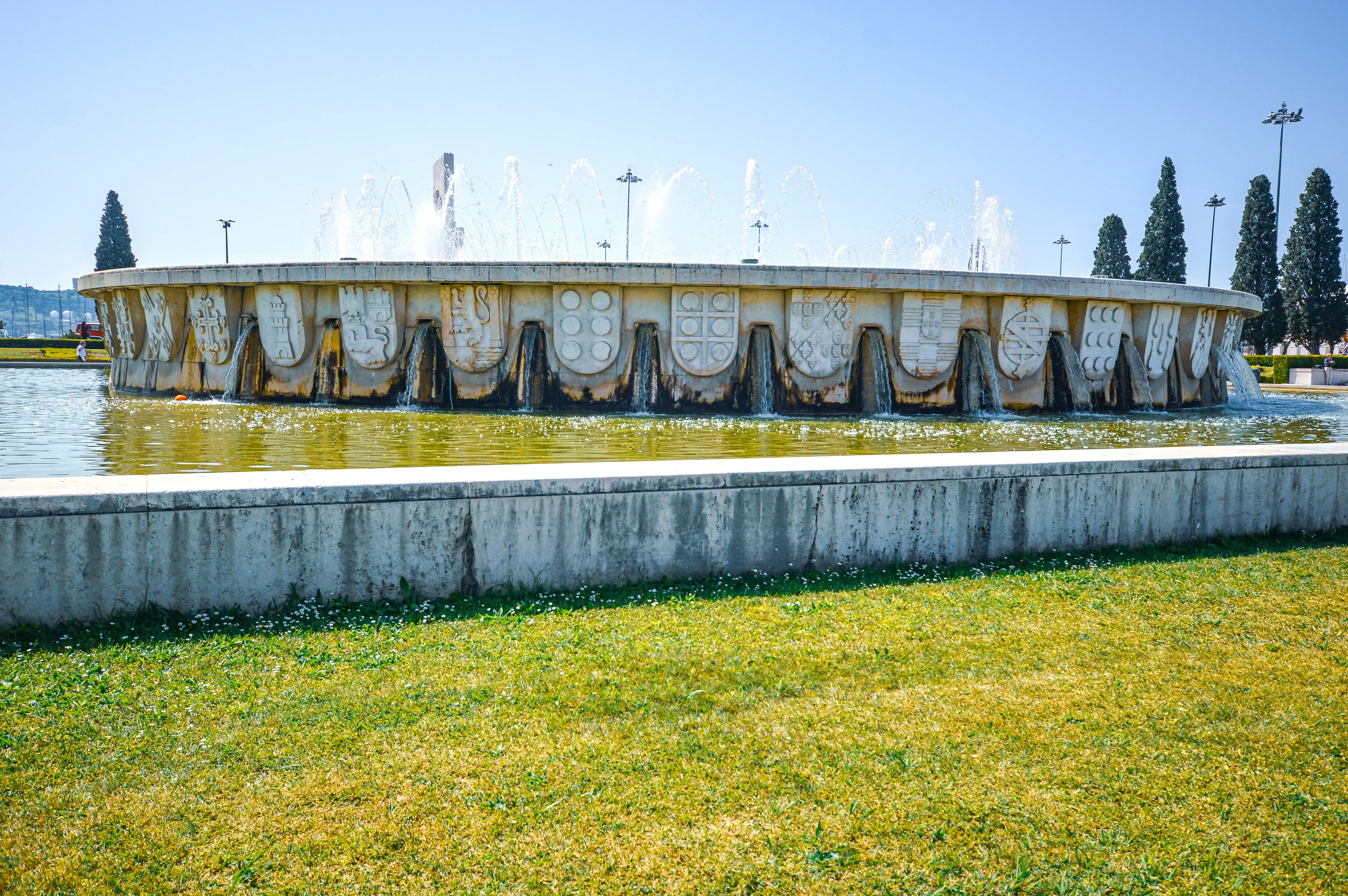A circular fountain with detailed stone carvings and flowing water, set against a backdrop of trees and a blue sky.